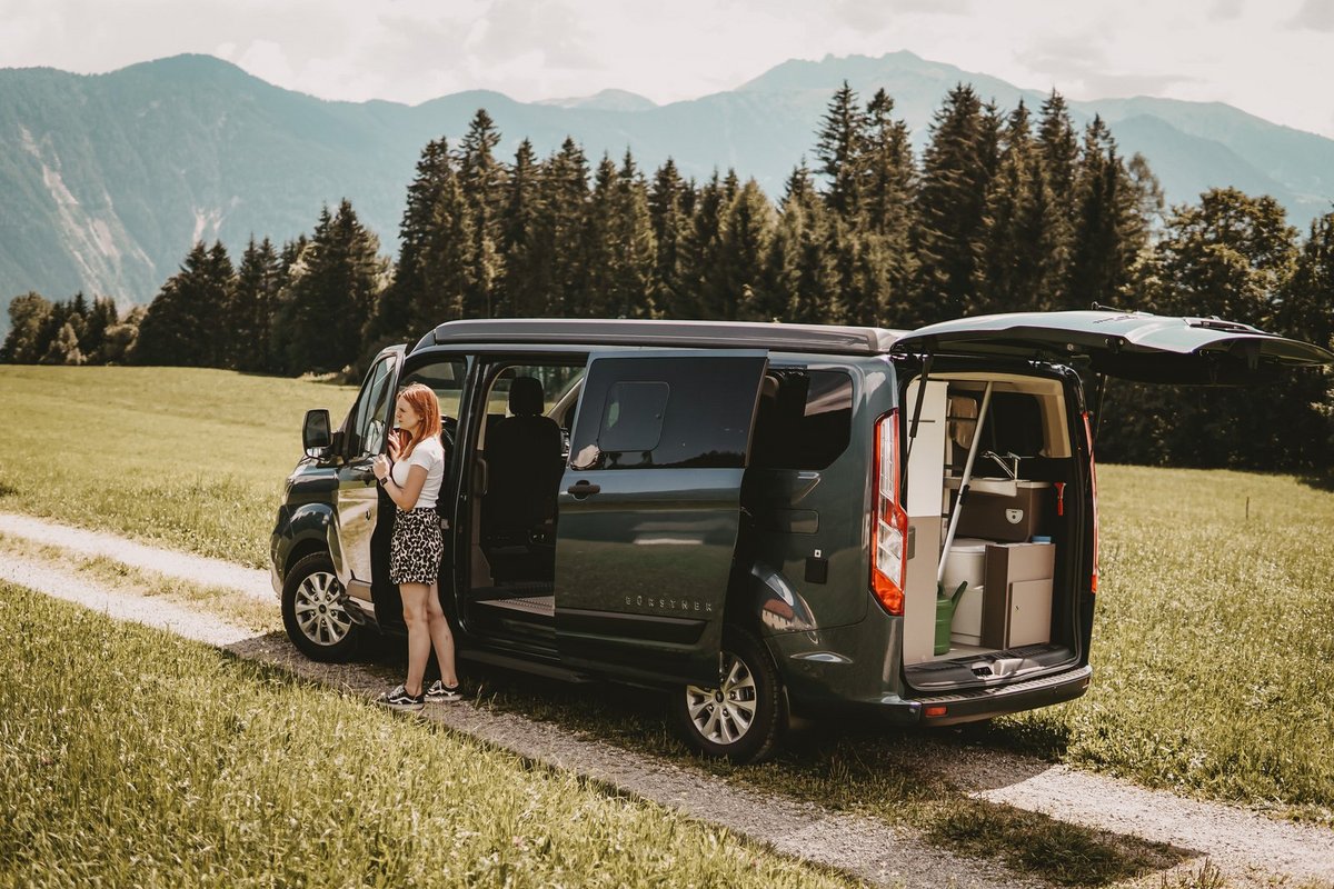 Frau steht neben Bürstner Van, Wald und Berge im Hintergrund